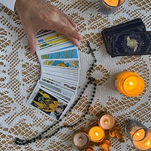 Puede incluir: Una configuraci&oacute;n de lectura de cartas del tarot con una baraja de cartas en abanico, un rosario y velas encendidas. Las cartas tienen ilustraciones coloridas y el texto "THE DEVIL" es visible en una carta. La escena est&aacute; ambientada sobre un mantel de encaje blanco.