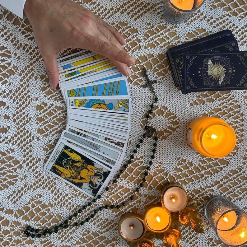 Puede incluir: Una configuraci&oacute;n de lectura de cartas del tarot con una baraja de cartas en abanico, un rosario y velas encendidas. Las cartas tienen ilustraciones coloridas y el texto "THE DEVIL" es visible en una carta. La escena est&aacute; ambientada sobre un mantel de encaje blanco.