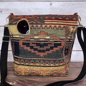 May include: A square crossbody bag with a woven tapestry design in shades of red, black, gold, and green. The bag has a black adjustable strap and a pair of sunglasses in the front pocket. The bag is sitting on a wooden surface.