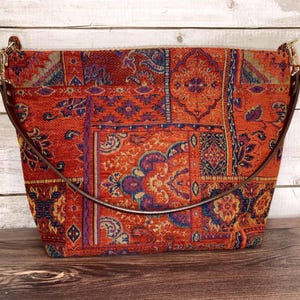May include: A structured handbag featuring a vibrant orange tapestry fabric with a multi-colored floral and geometric pattern. The bag includes a dark brown leather strap attached with gold-toned metal hardware on both sides.