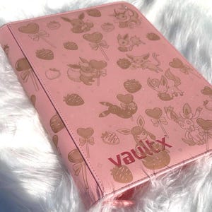 May include: A pink binder featuring a pattern of cartoon characters, strawberries, and heart-shaped lollipops. The word "Vaultx" is printed in reddish-brown. The binder is closed and has a zip closure.