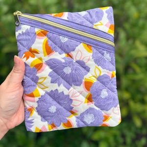 May include: Quilted clutch with a floral pattern in purple, orange, yellow, and pink on a white background. The clutch has a gold zipper and a lavender border. The bag is held in a hand against a green background.