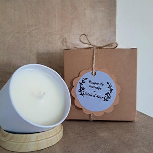 Peut inclure: Une bougie blanche dans un récipient bleu clair repose sur une base en bois. Un coffret cadeau marron avec une étiquette indiquant "Bougie de massage Soleil d'Azur" est noué avec de la ficelle. La bougie est non parfumée.
