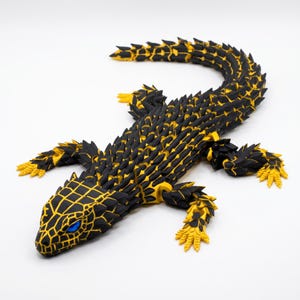 Puede incluir: Una figura de lagarto articulada con un patrón geométrico de escamas negras y amarillas en todo el cuerpo. La criatura tiene una cola larga segmentada, cuatro patas detalladas con garras y ojos de color azul brillante en una cabeza texturizada.