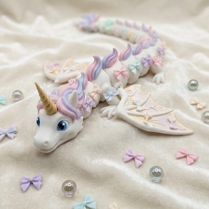 May include: A white articulated dragon figurine featuring a gold horn, blue eyes, and a mane of pastel pink, purple, and blue. The body is adorned with small pastel bows, and the wings display a gradient pattern. Scattered small bows and beads surround it.
