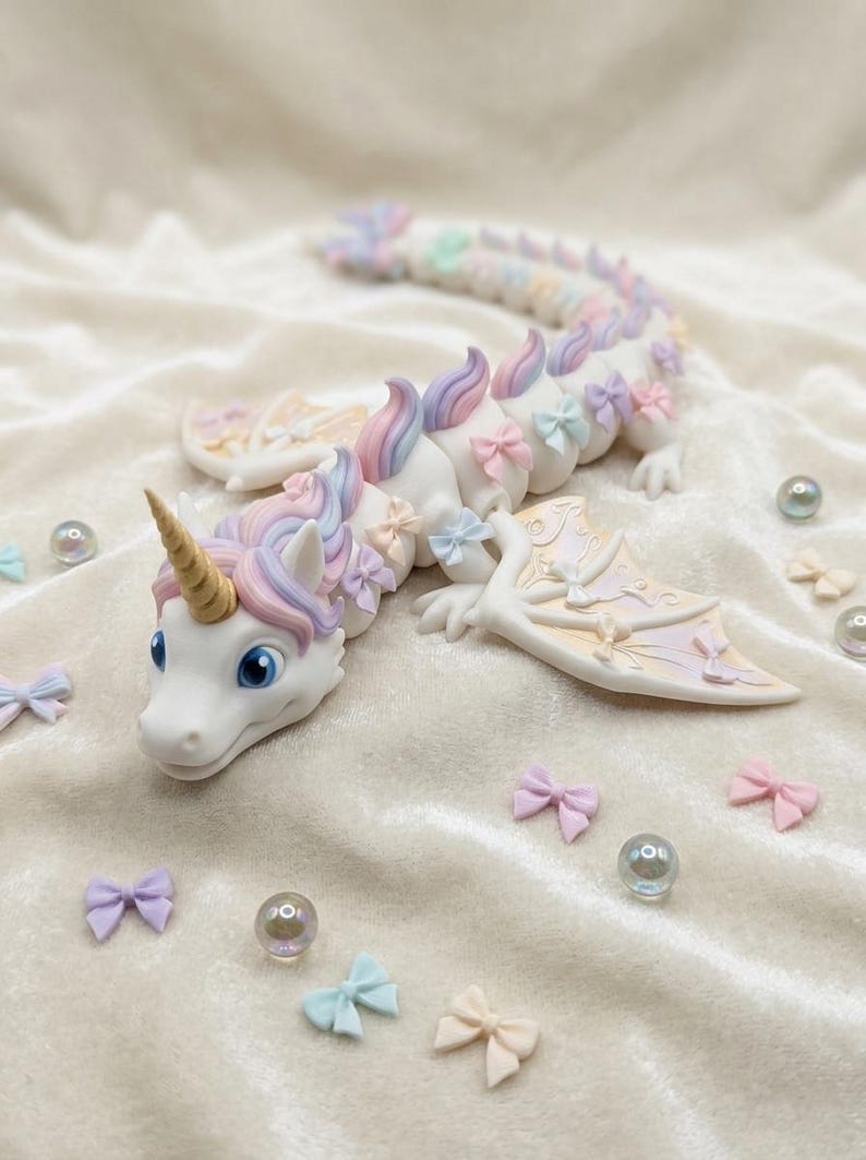 May include: A white articulated dragon figurine featuring a gold horn, blue eyes, and a mane of pastel pink, purple, and blue. The body is adorned with small pastel bows, and the wings display a gradient pattern. Scattered small bows and beads surround it.