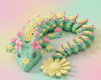 Puede incluir: Una figura de dragón articulada de color verde menta con una superficie texturizada similar a escamas. La criatura está adornada con numerosas margaritas amarillas pequeñas y capullos de flores rosas en el lomo y la cabeza. Una margarita amarilla grande descansa al frente.