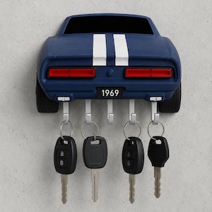 May include: A dark blue wall-mounted key holder shaped like the rear of a 1969 muscle car, featuring two white racing stripes, red taillights, and a license plate reading 1969. It includes five silver metal hooks, four of which hold black car keys.