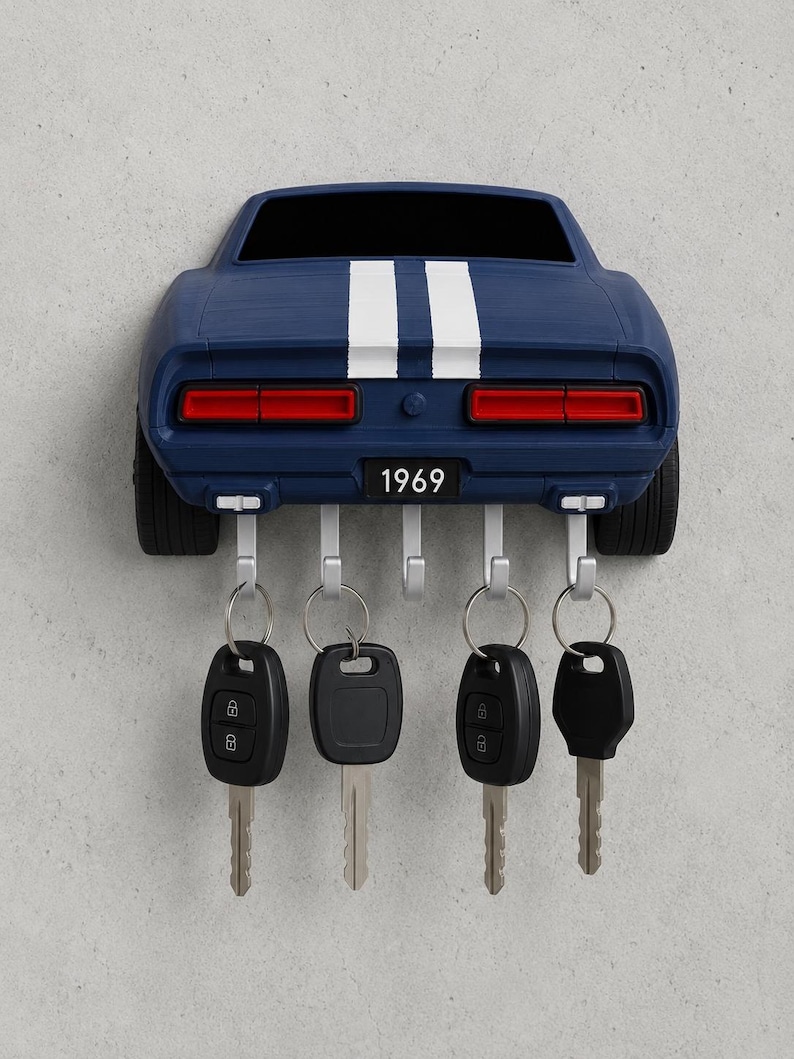 May include: A dark blue wall-mounted key holder shaped like the rear of a 1969 muscle car, featuring two white racing stripes, red taillights, and a license plate reading 1969. It includes five silver metal hooks, four of which hold black car keys.