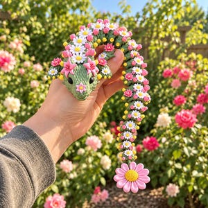 May include: A green articulated lizard figurine featuring a textured scale pattern, decorated with numerous small white daisies, pink flowers, and yellow accents along its back and tail, ending with a large pink daisy at the tip of the tail.