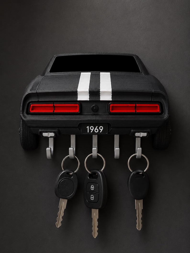 May include: A black wall-mounted key holder shaped like the rear of a 1969 muscle car. It features two white racing stripes, red taillights, and a license plate reading 1969. Five silver hooks extend from the bottom, holding three metal key rings.