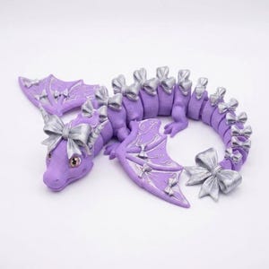 May include: A lavender articulated dragon figurine featuring metallic silver bows attached to its head, wings, and along its segmented tail. The dragon has large, light-colored eyes and intricate silver line patterns on its purple wings.