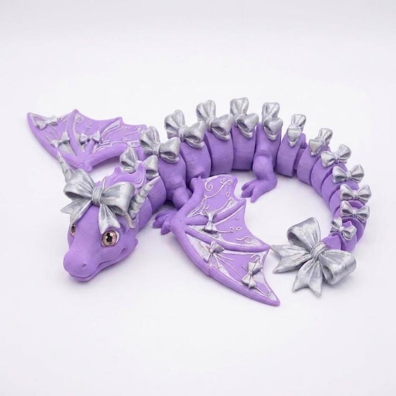 May include: A lavender articulated dragon figurine featuring metallic silver bows attached to its head, wings, and along its segmented tail. The dragon has large, light-colored eyes and intricate silver line patterns on its purple wings.