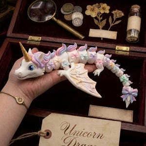 May include: A white articulated dragon figurine with a gold horn, pastel-colored mane, and small bows along its body. The dragon features wings and a long, segmented tail. A beige tag attached to the figure displays the text Unicorn Dragon.