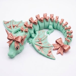 May include: A mint green articulated dragon figurine featuring metallic rose gold bows attached to its head, wings, and along the length of its segmented body and tail. The dragon has large eyes and a curved posture on a plain white background.