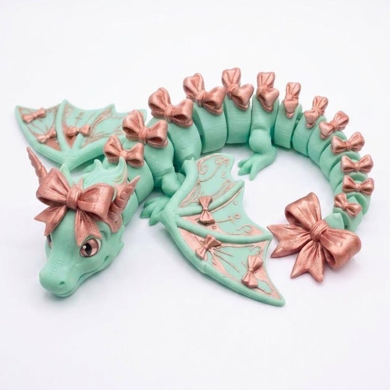 May include: A mint green articulated dragon figurine featuring metallic rose gold bows attached to its head, wings, and along the length of its segmented body and tail. The dragon has large eyes and a curved posture on a plain white background.