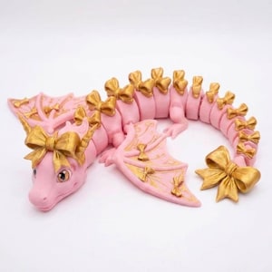 May include: A pink articulated dragon figurine featuring gold-colored bows attached to its head, wings, and along the length of its segmented body. The dragon has large eyes and a curved tail ending in a prominent gold bow, with gold accents on wings.