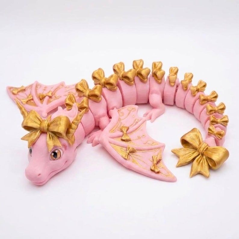 May include: A pink articulated dragon figurine featuring gold-colored bows attached to its head, wings, and along the length of its segmented body. The dragon has large eyes and a curved tail ending in a prominent gold bow, with gold accents on wings.