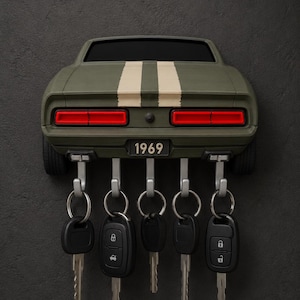 May include: A wall-mounted key holder shaped like the rear of a matte olive green 1969 muscle car with two white racing stripes. The vehicle features red taillights and a license plate reading 1969. Five silver hooks hold five black car keys.