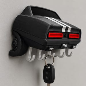 May include: A black wall-mounted key holder shaped like the rear of a 1969 muscle car. The car features white racing stripes, red taillights, and a license plate reading 1969. Four silver hooks extend from the bottom, with one key hanging.