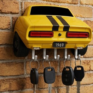 May include: A yellow wall-mounted key holder shaped like the rear of a 1969 muscle car with two black racing stripes. The car model features red taillights, black tires on the sides, and a license plate reading 1969. Five silver hooks hold four key rings.