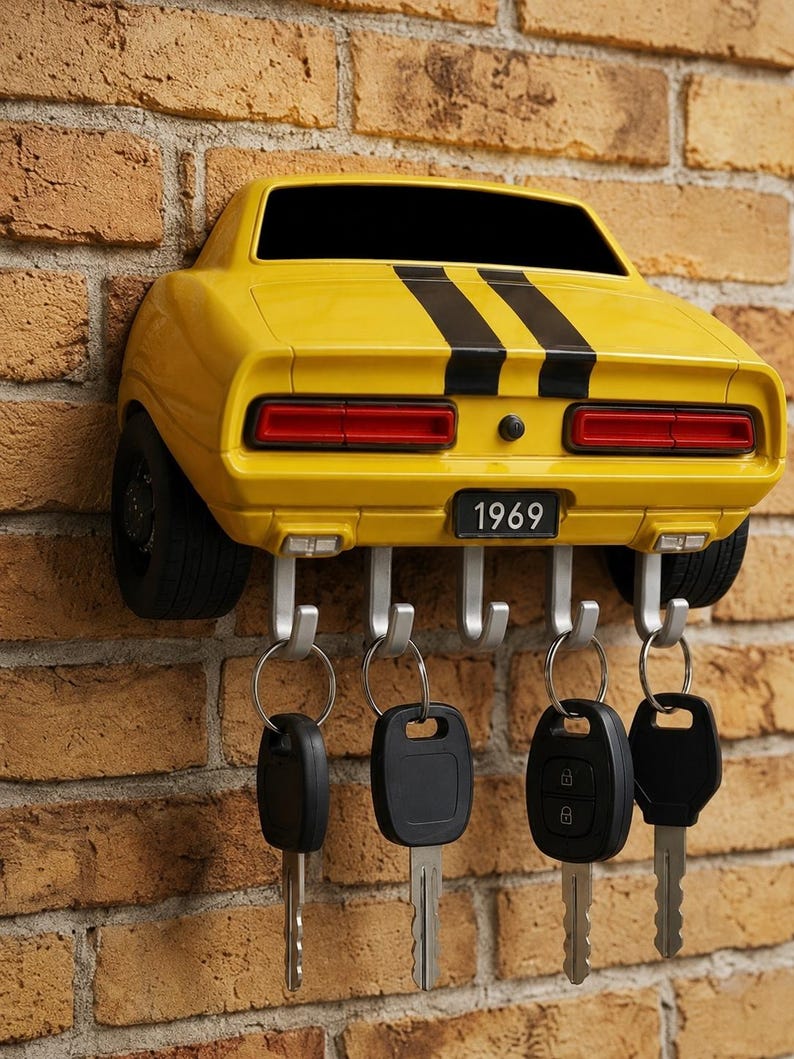 May include: A yellow wall-mounted key holder shaped like the rear of a 1969 muscle car with two black racing stripes. The car model features red taillights, black tires on the sides, and a license plate reading 1969. Five silver hooks hold four key rings.