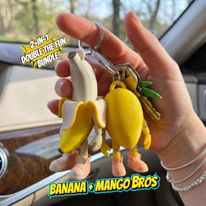 Op de afbeelding: Een sleutelhangerbundel met een banaan- en mangokarakter. De banaan is geschild en onthult een wit interieur, en de mango is geel. Beide hebben armen, benen en schoenen. De tekst "2-IN-1 DOUBLE THE FUN BUNDLE" en "BANANA + MANGO BROS" is ook zichtbaar.
