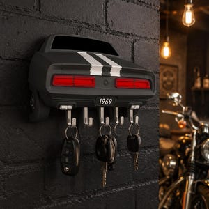 May include: A wall-mounted key holder shaped like the rear of a black 1969 muscle car. It features two white racing stripes, red taillights, and a license plate reading 1969. Five silver metal hooks extend from the bottom, holding various keys.