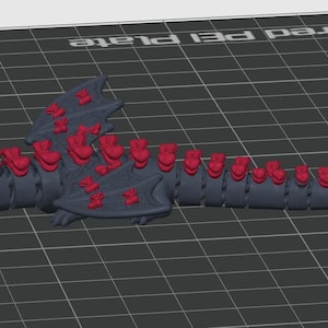 May include: A dark gray articulated dragon figurine featuring a series of bright red bows attached along its back, wings, and tail, with a large red bow positioned on its head and another at the tip of its tail, all set against a grid background.