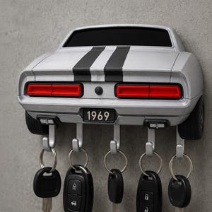 May include: A silver wall-mounted key holder shaped like the rear of a 1969 muscle car. It features two black racing stripes, red taillights, and a license plate reading 1969. Five silver hooks extend from the bottom, each holding a set of keys.