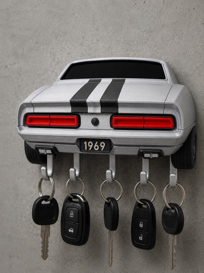 May include: A silver wall-mounted key holder shaped like the rear of a 1969 muscle car. It features two black racing stripes, red taillights, and a license plate reading 1969. Five silver hooks extend from the bottom, each holding a set of keys.