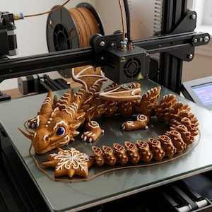 May include: A brown, textured dragon figurine with white icing details and blue eyes, positioned on the glass print bed of a black 3D printer. A brown snowflake-shaped cookie with white icing sits in front of the dragon, near the printer nozzle.