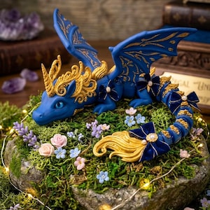 May include: A blue dragon figurine with gold filigree details on its wings and head, featuring a spiraled gold horn, blue velvet bows with pearl centers on its body and tail, and a flowing gold tail tip, resting on a mossy surface with flowers.