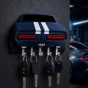 May include: A wall-mounted key holder shaped like the rear of a dark blue 1969 muscle car with two white racing stripes. The design features red taillights and a license plate reading 1969. Five silver metal hooks hold four black car key fobs.