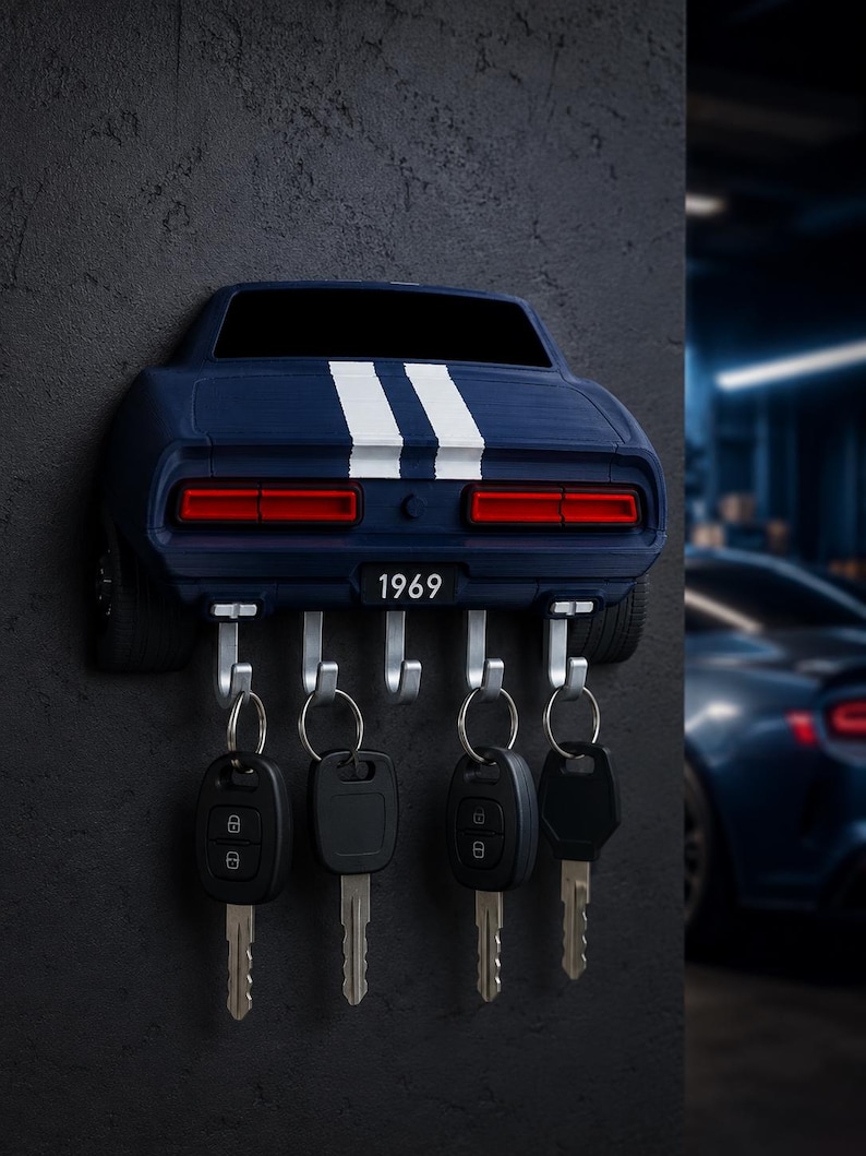 May include: A wall-mounted key holder shaped like the rear of a dark blue 1969 muscle car with two white racing stripes. The design features red taillights and a license plate reading 1969. Five silver metal hooks hold four black car key fobs.