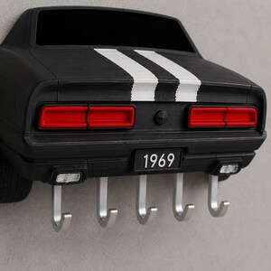 May include: A black wall-mounted key holder shaped like the rear of a 1969 muscle car. It features two white racing stripes, red taillights, and a license plate reading 1969. Five silver metal hooks extend from the bottom for hanging items.