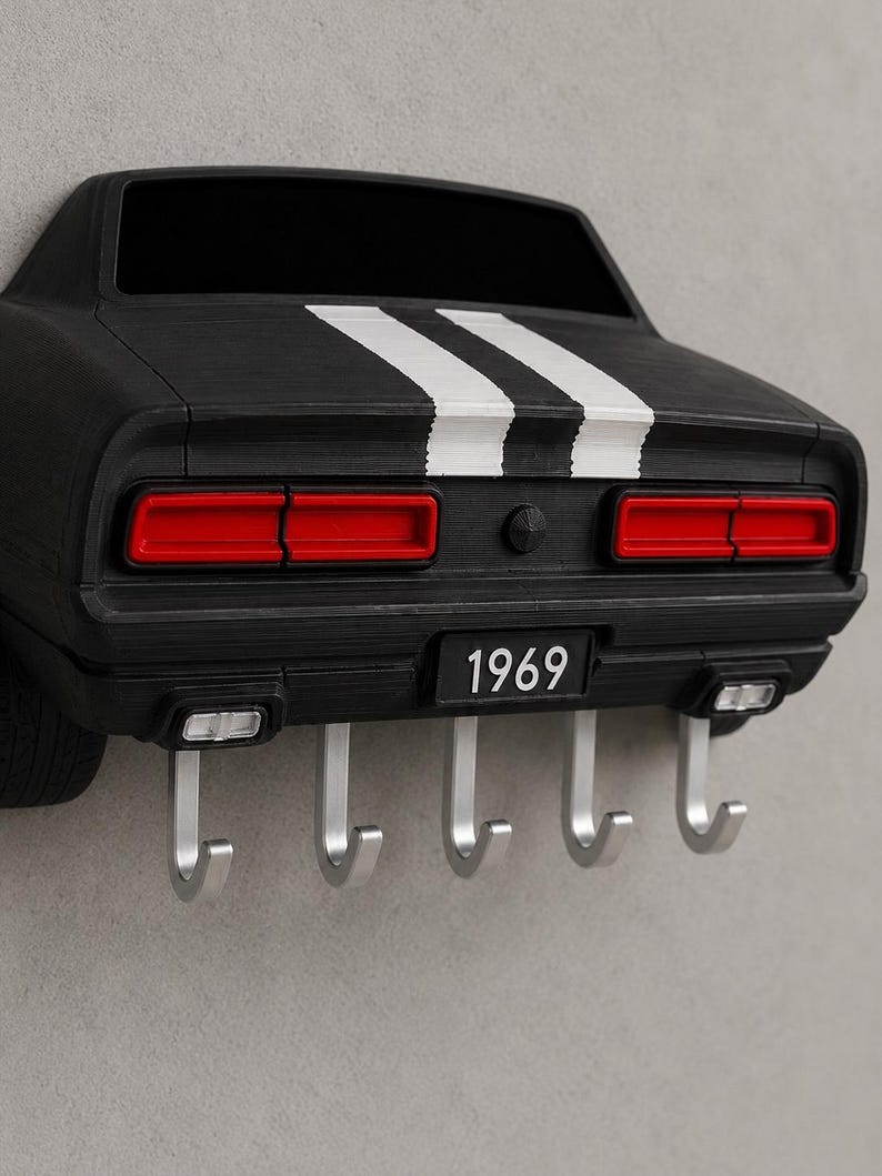 May include: A black wall-mounted key holder shaped like the rear of a 1969 muscle car. It features two white racing stripes, red taillights, and a license plate reading 1969. Five silver metal hooks extend from the bottom for hanging items.