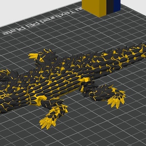 May include: A 3D printed articulated lizard figure featuring a black and yellow scale pattern with bright blue eyes. The lizard is positioned on a dark gray grid-patterned build plate with the text Textured PEI Plate visible in the corner.