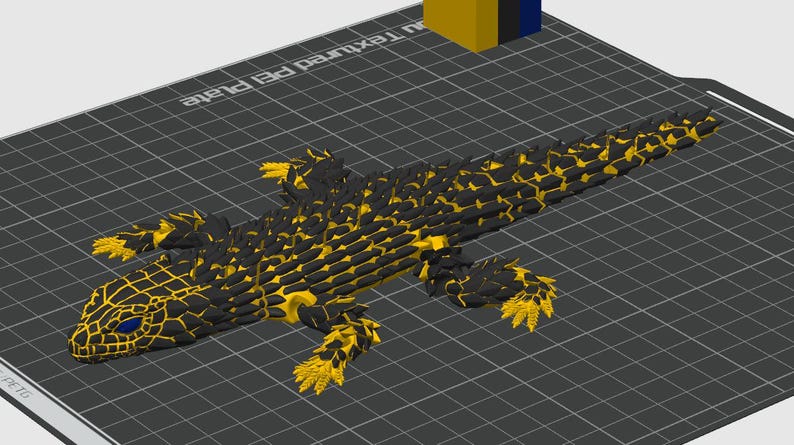May include: A 3D printed articulated lizard figure featuring a black and yellow scale pattern with bright blue eyes. The lizard is positioned on a dark gray grid-patterned build plate with the text Textured PEI Plate visible in the corner.