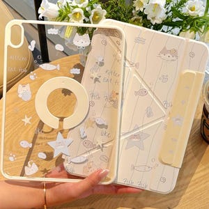 May include: A beige and clear tablet case featuring a cat and fish design. The clear case has a ring holder and the text "Kittens Eat Fish". The case is open, revealing the interior design. The brand name "CuteXCaseDesign" is visible.