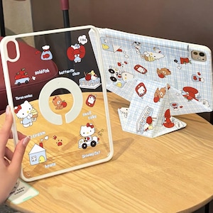 Könnte beinhalten: Transparente Tablet-Hülle mit weißem Rahmen, mit farbenfrohen Cartoon-Grafiken von Hello Kitty, Bären und anderen niedlichen Illustrationen. Auf der Hülle stehen die Wörter "goldfish", "butterfly clasp", "small cake", "Driving Cat" und "I like". Das Tablet liegt auf einem Holztisch.