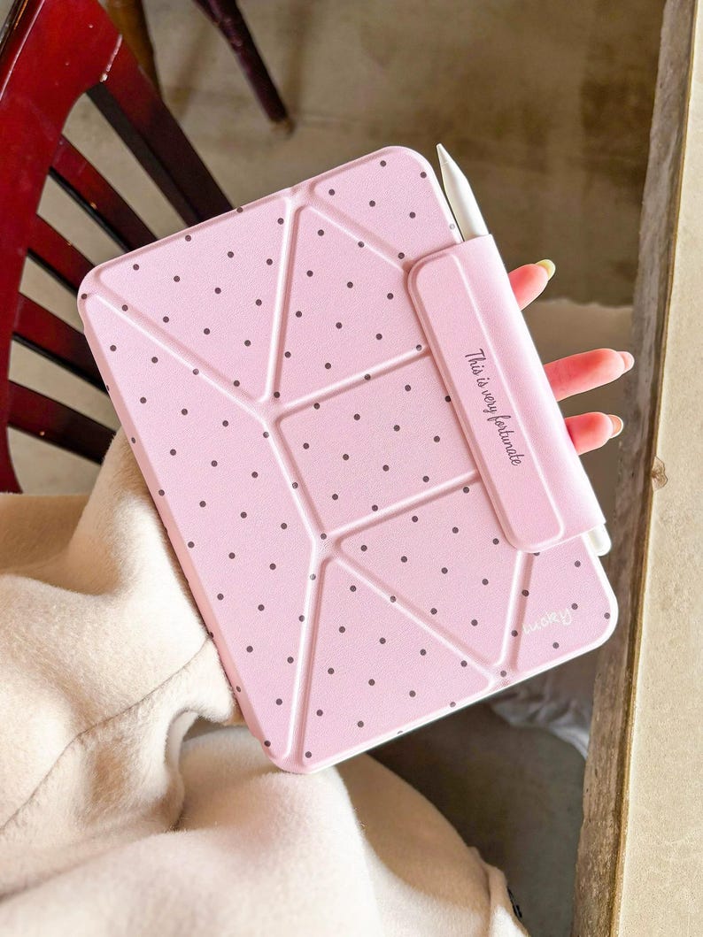 Op de afbeelding: Een lichtroze tablethoes met een geometrisch ontwerp en kleine zwarte stippen. De hoes heeft een ingebouwde houder voor een witte stylus. De tekst "This is very fortunate" staat op de stylus houder.