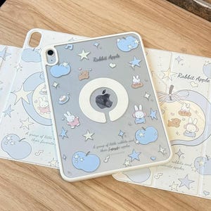 Op de afbeelding: Een lichtgrijze iPad-hoes met een witte rand, met een cartoonontwerp van konijntjes, sterren en appels. De tekst "Rabbit Apple" is zichtbaar. De hoes wordt getoond met een bijpassende hoes.