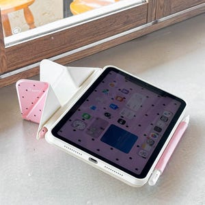 Op de afbeelding: Een iPad in een witte hoes met een roze stylus en een roze standaard met aardbeienpatroon. Het iPad-scherm toont een roze achtergrond met zwarte stericonen. De opstelling staat op een wit oppervlak bij een raam.