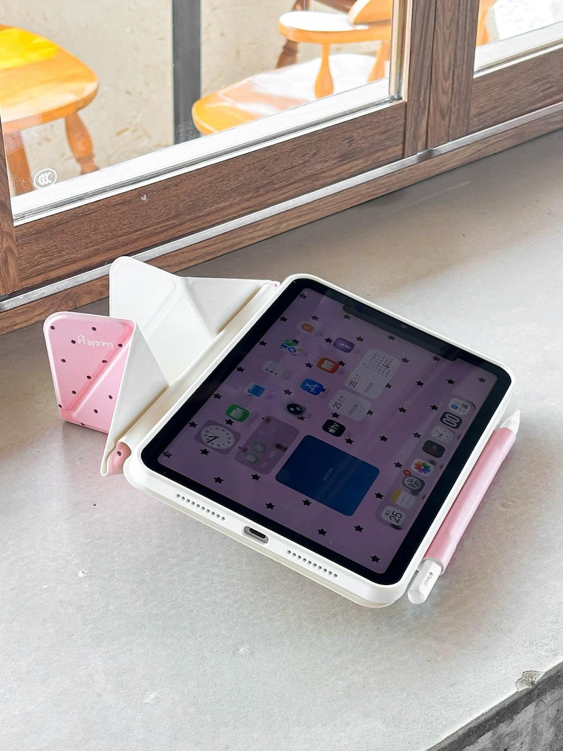 Op de afbeelding: Een iPad in een witte hoes met een roze stylus en een roze standaard met aardbeienpatroon. Het iPad-scherm toont een roze achtergrond met zwarte stericonen. De opstelling staat op een wit oppervlak bij een raam.