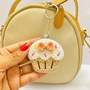 Może przedstawiać: Szydełkowy brelok do kluczy w kształcie babeczki z białym wierzchem, beżową podstawą i brzoskwiniową kokardką. Wierzch ma różowe i żółte posypki. Brelok jest przymocowany do srebrnego pierścienia i beżowej torebki.