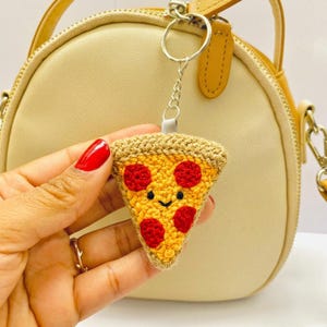 Może przedstawiać: Szydełkowy brelok do kluczy w kształcie kawałka pizzy z uśmiechniętą buzią i czerwonym pepperoni. Brelok jest przymocowany do srebrnego łańcuszka i kółka. Kawałek pizzy jest żółty i brązowy.