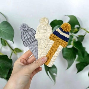 Puede incluir: Tres adornos de gorros de invierno de punto en gris, crema y amarillo mostaza con pompones. Los adornos se sostienen en una mano sobre un fondo de hojas verdes. Los gorros tienen diferentes patrones de punto y combinaciones de colores.