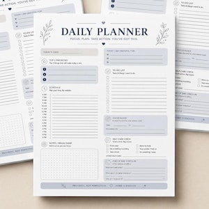 Puede incluir: Tres blocs de notas Daily Planner blancos con texto azul y detalles florales. Cada bloc de notas presenta secciones para tareas diarias, prioridades y notas. El texto dice "Daily Planner: Focus, Plan, Take Action, You've Got This."