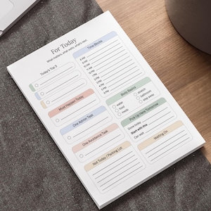 May include: A white notepad with the title "For Today" and sections for tasks, time blocks, and body basics. The notepad is on a wooden surface next to a laptop and a potted plant.